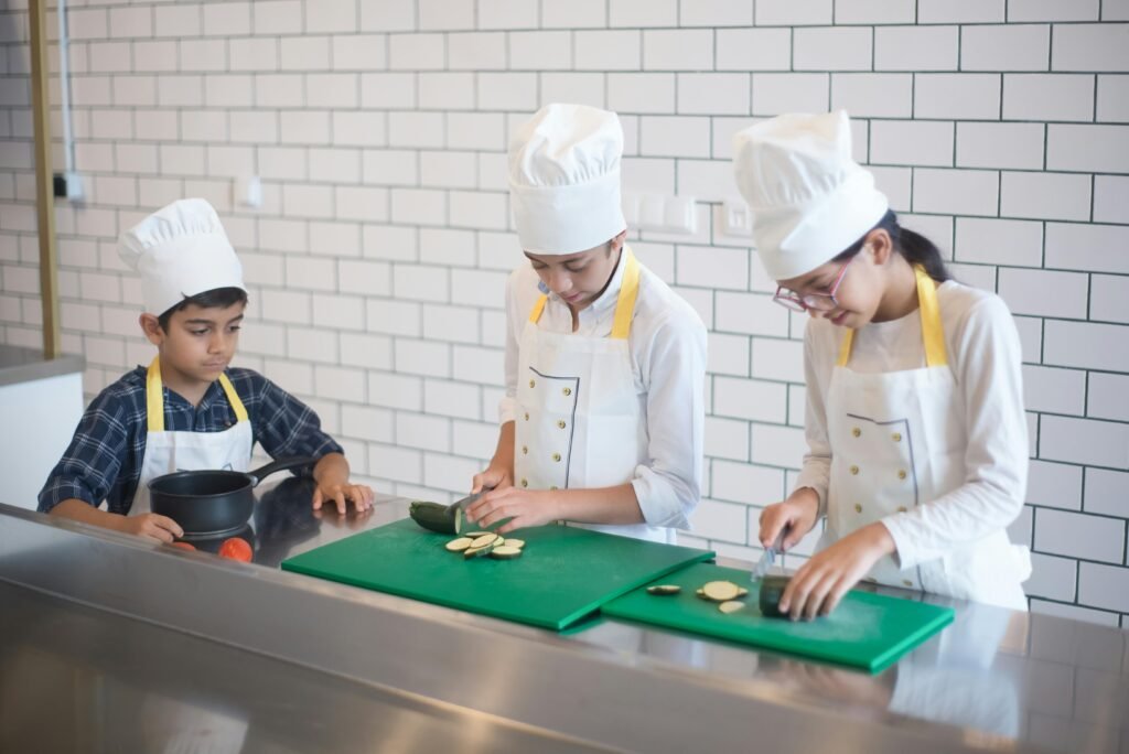 Kids wearing chef hats and aprons cooking in a professional kitchen setting.